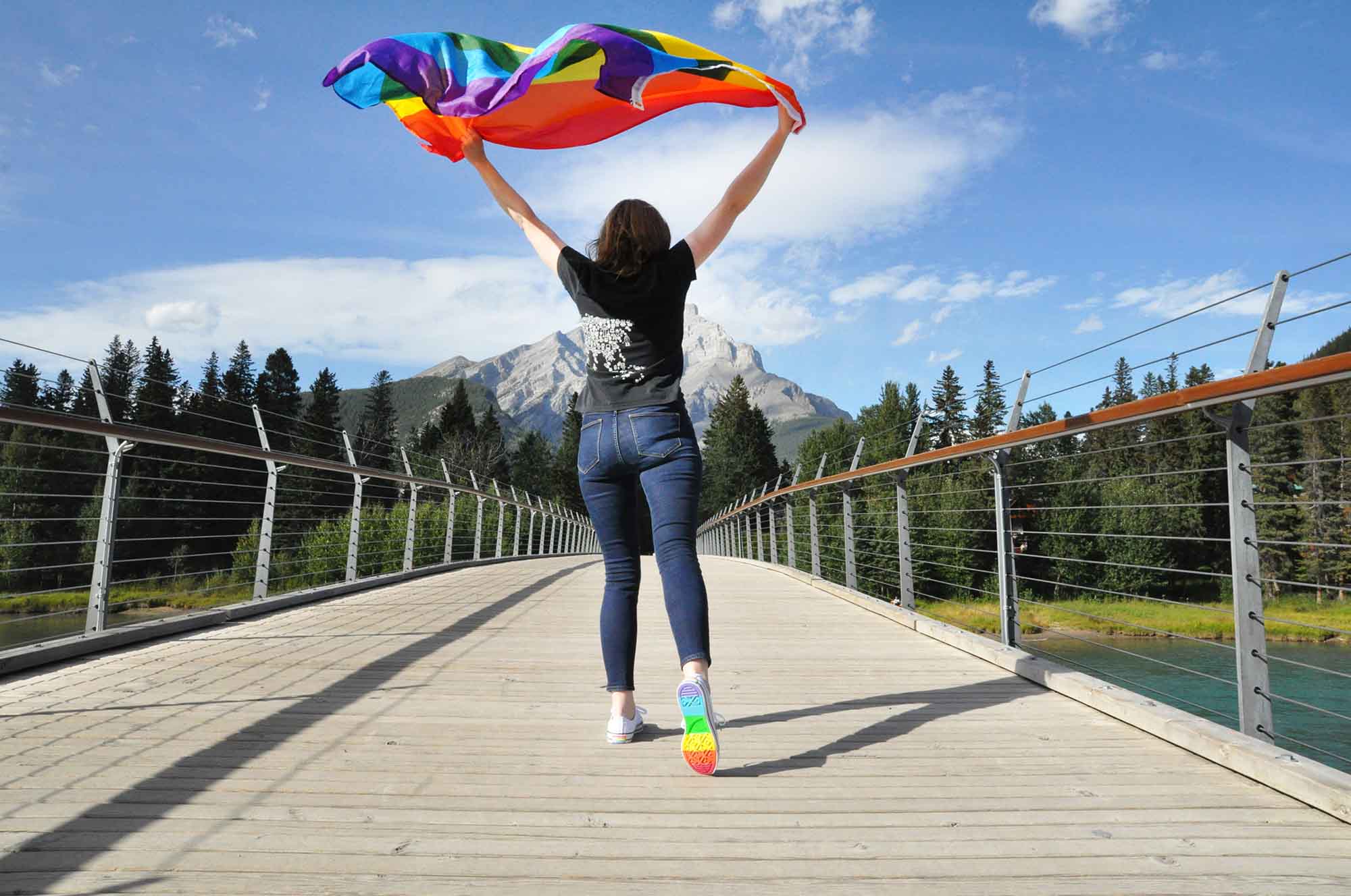 A person with a rainbow flag and rainbow shoes on the Banff Pedestrian Bridge for Banff Pride