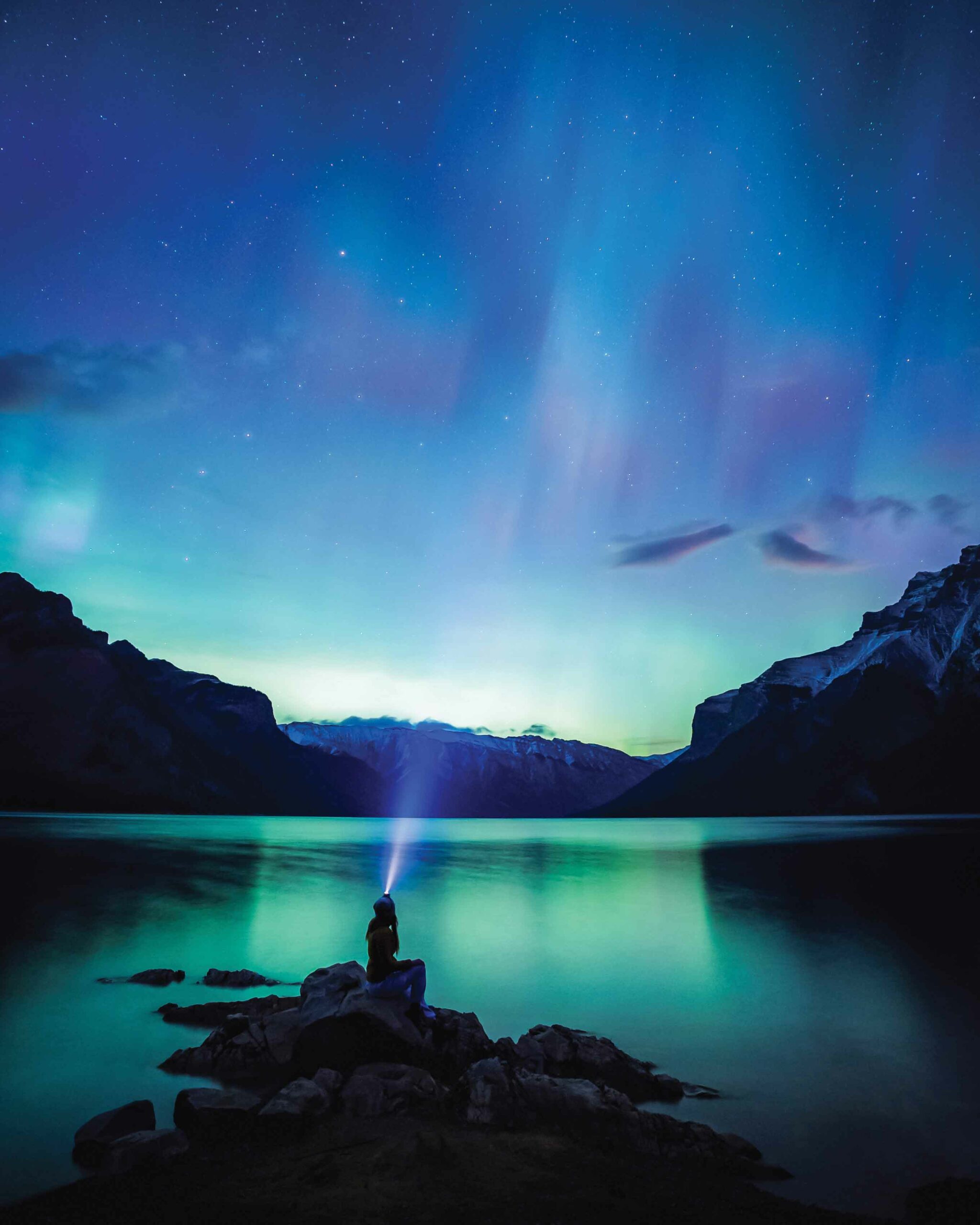 A person wearing a headlamp looks up at the northern lights reflected on the water.