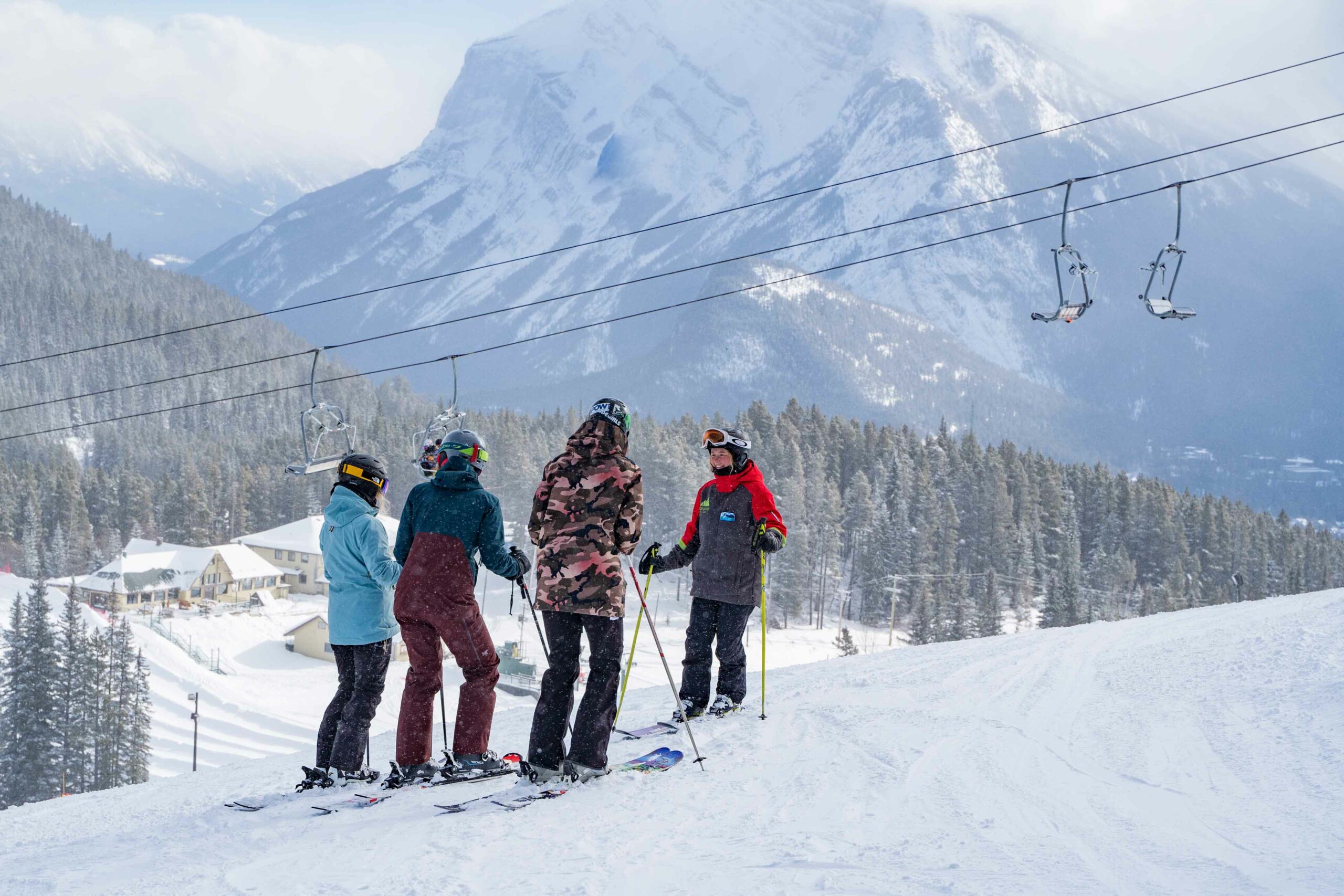 Three people stand facing a ski instructor on the hill at Mount Norquay with a mountain view in the background. 