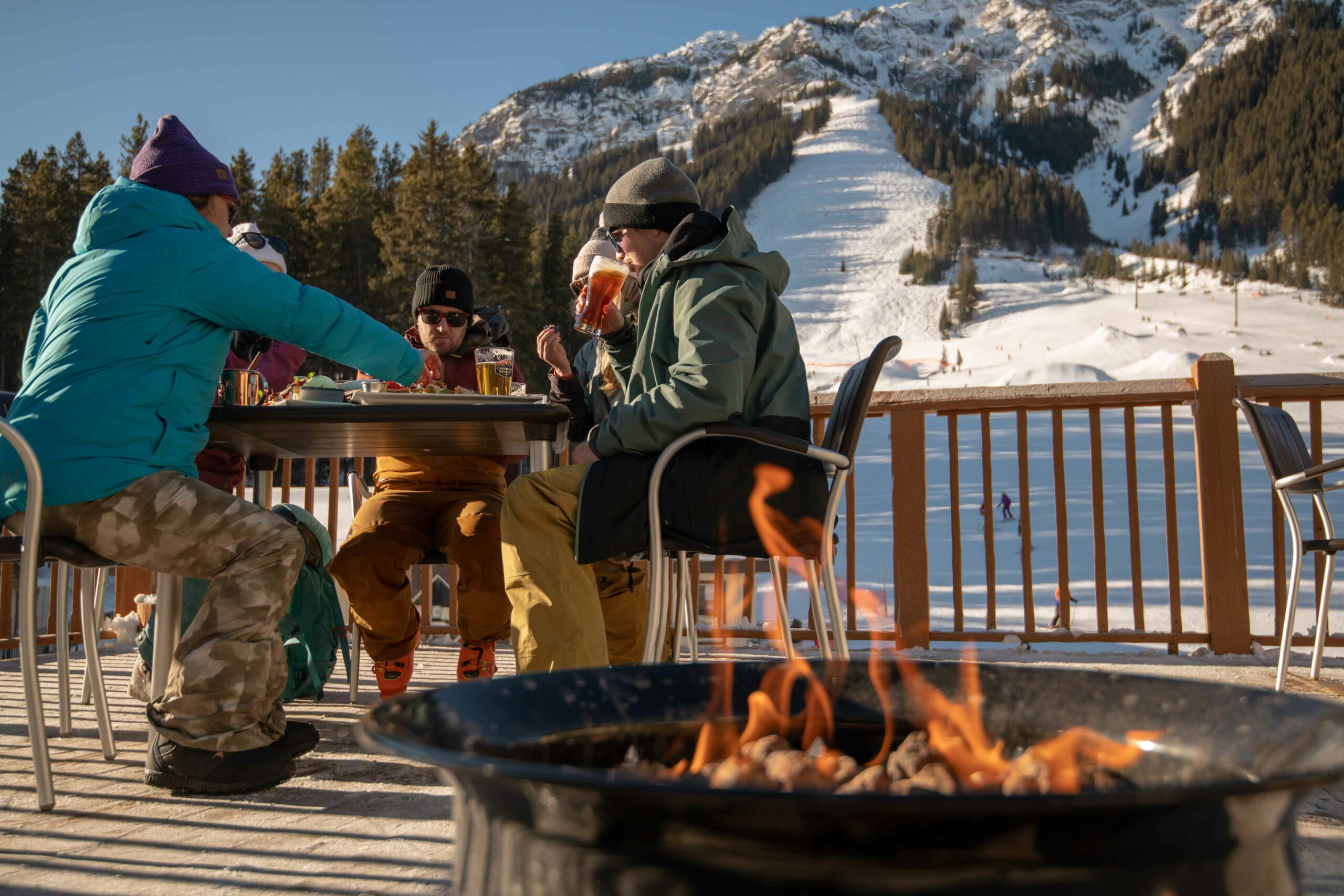 A group of friends enjoy food and drinks by the fireplace on the patio after skiing at Mount Norquay 