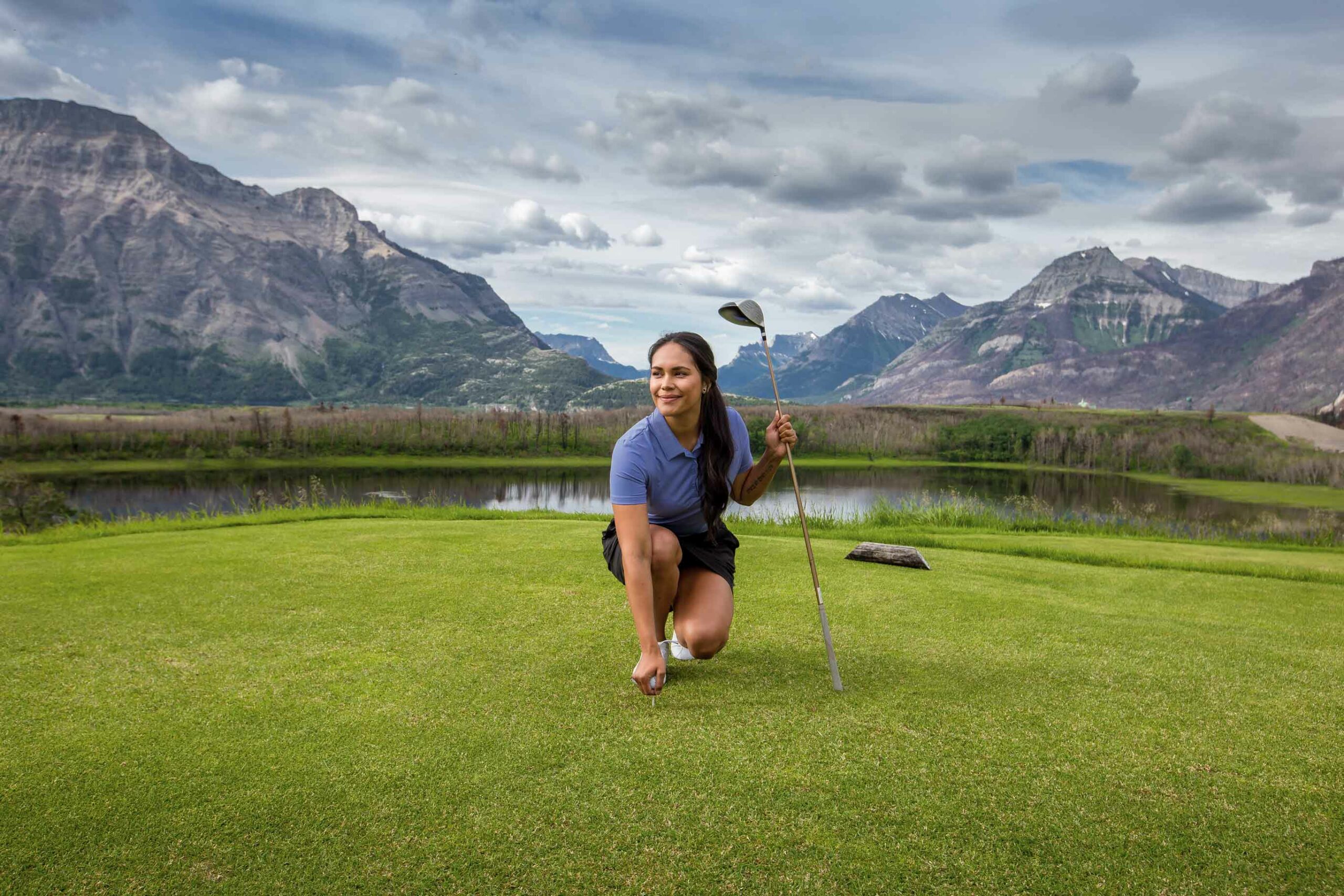 A woman kneels at on the green at the Waterton Lakes Golf Club with the mountain view in the background.