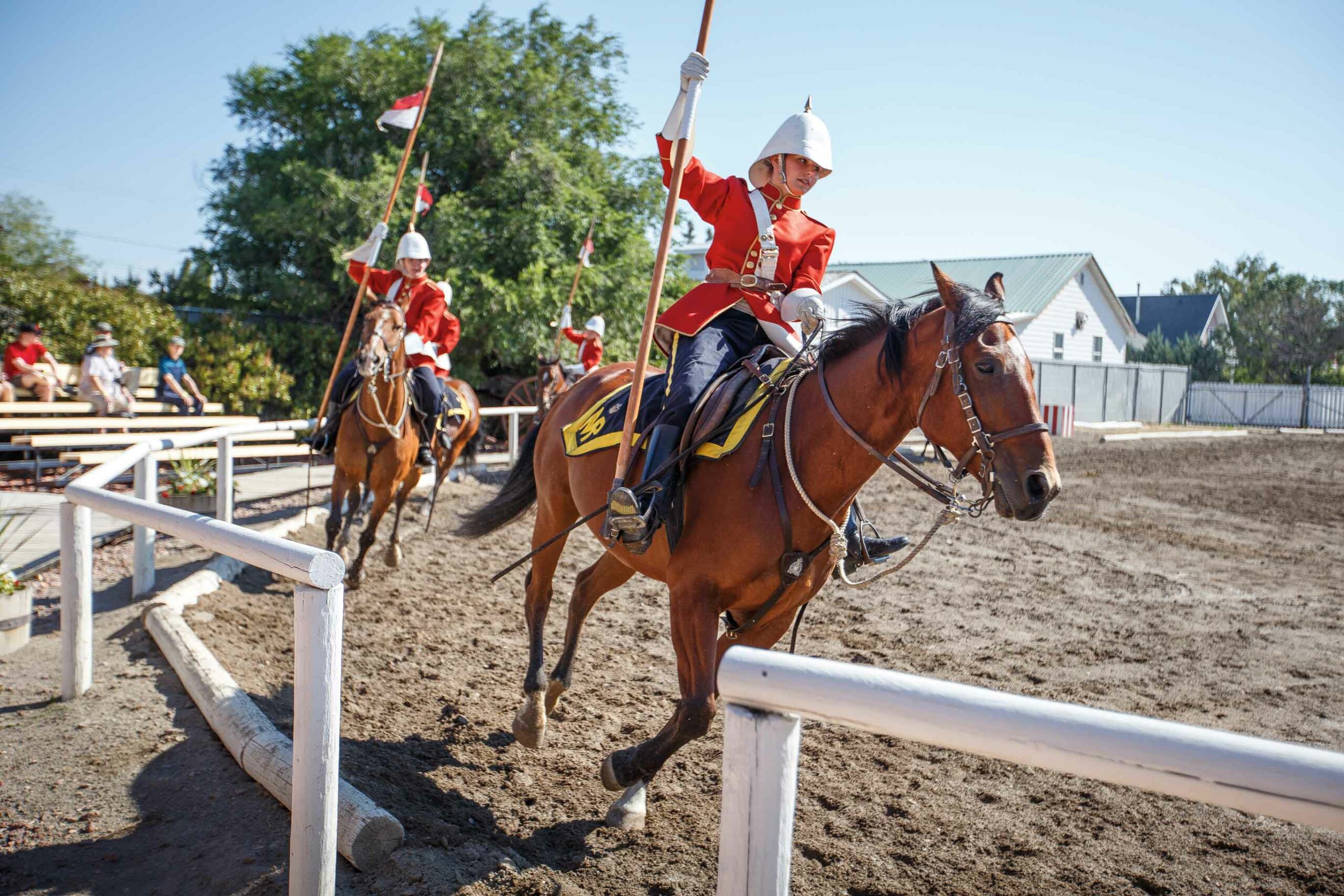 Red coats of the North West Mounted Police (NWMP) riding horses and carrying flags in outdoor arena in Southwestern Alberta.