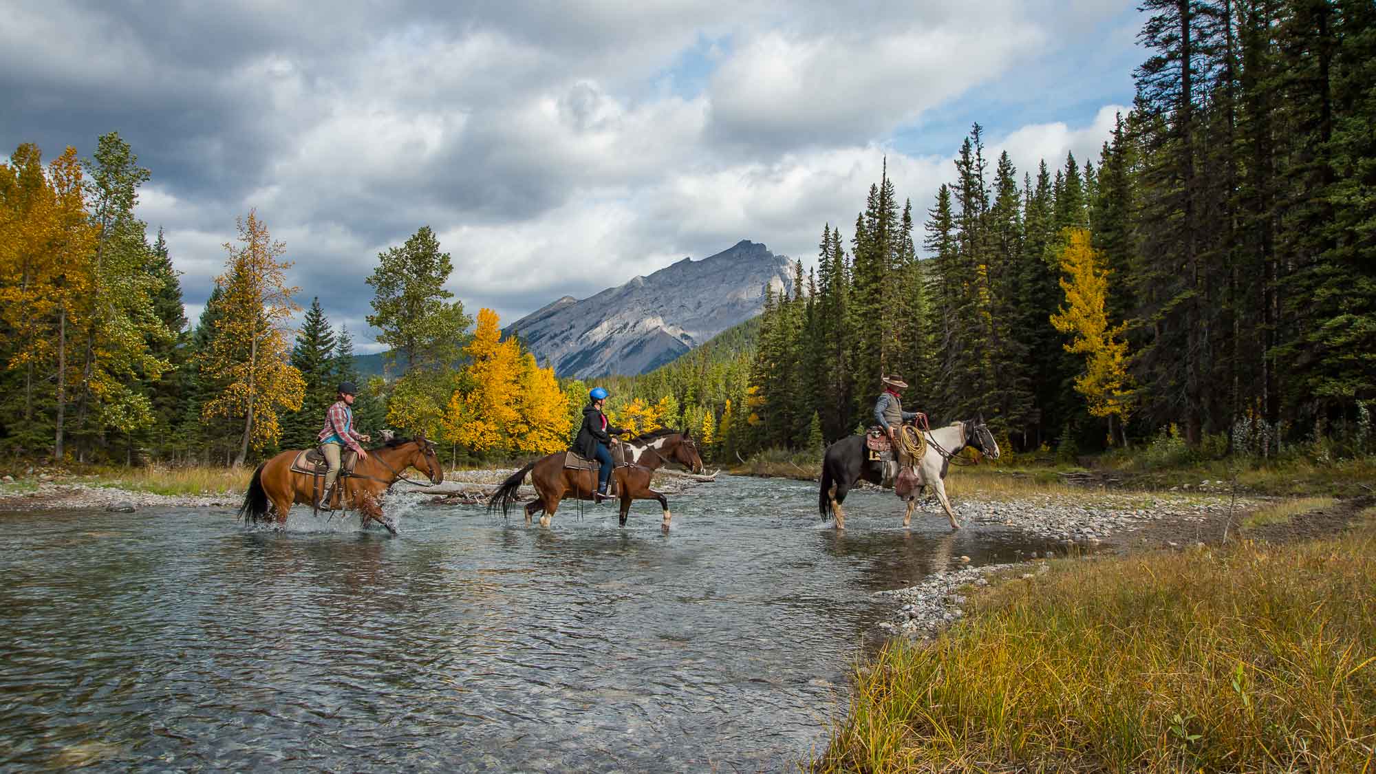 Three people live out their cowboy dreams and cross the river with mountains in the background on a trail ride with Banff Trail Riders. 